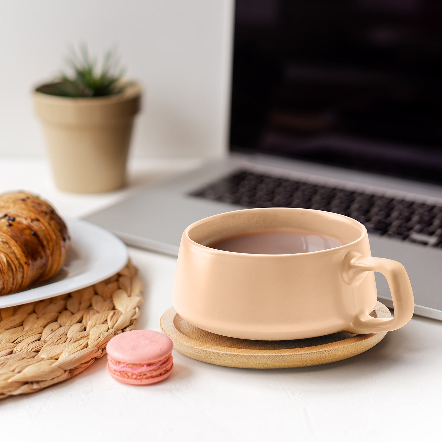 Tasse mit Kaffee auf einem Untersetzer aus Holz neben einem Croissant und Macaron, die vor einem Laptop steht