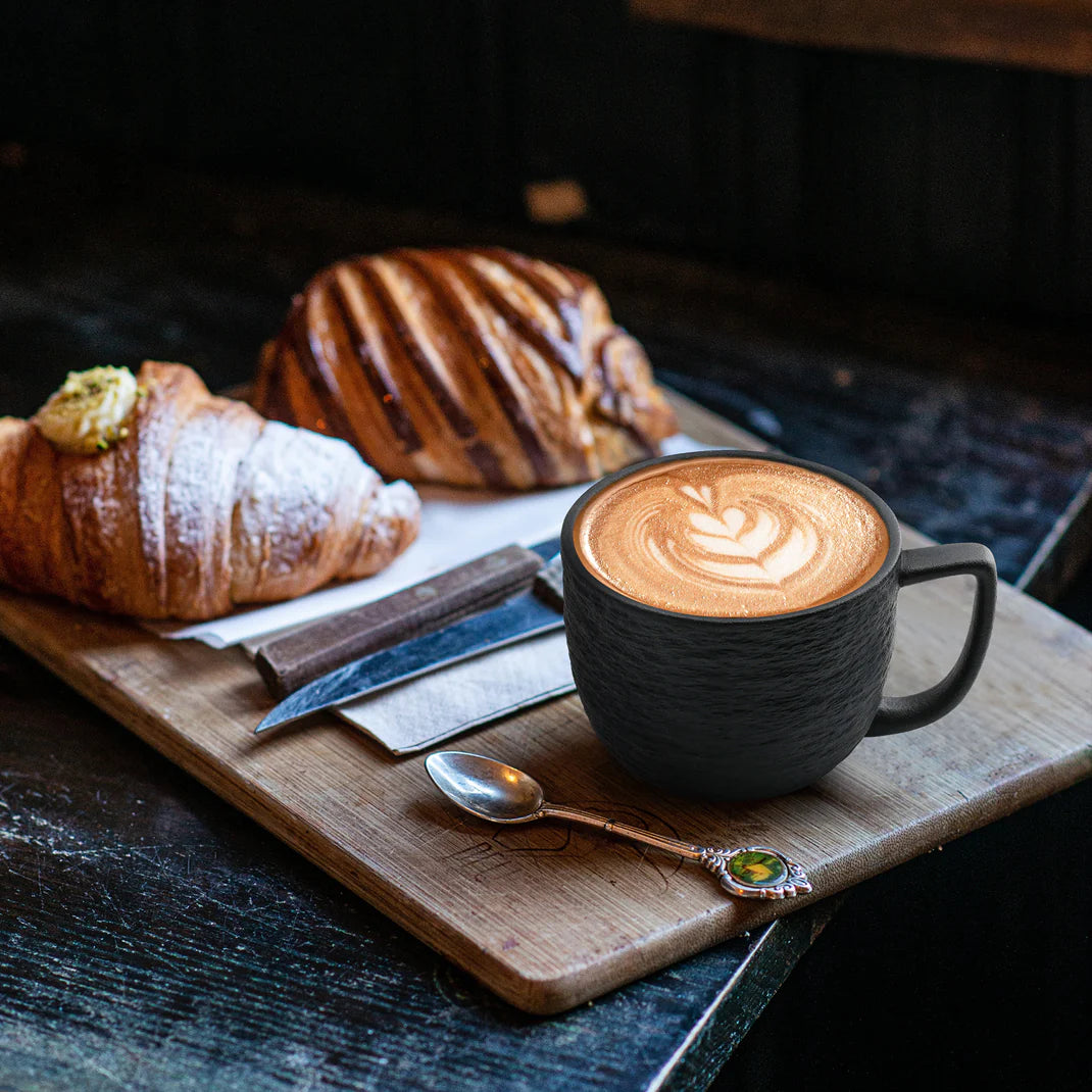 Schön angerichteter Cappuccino in einer MIAMIO Kaffeetasse der Oasis Kollektion auf einem Holzbrett mit Löffel neben verschiedenen Backwaren